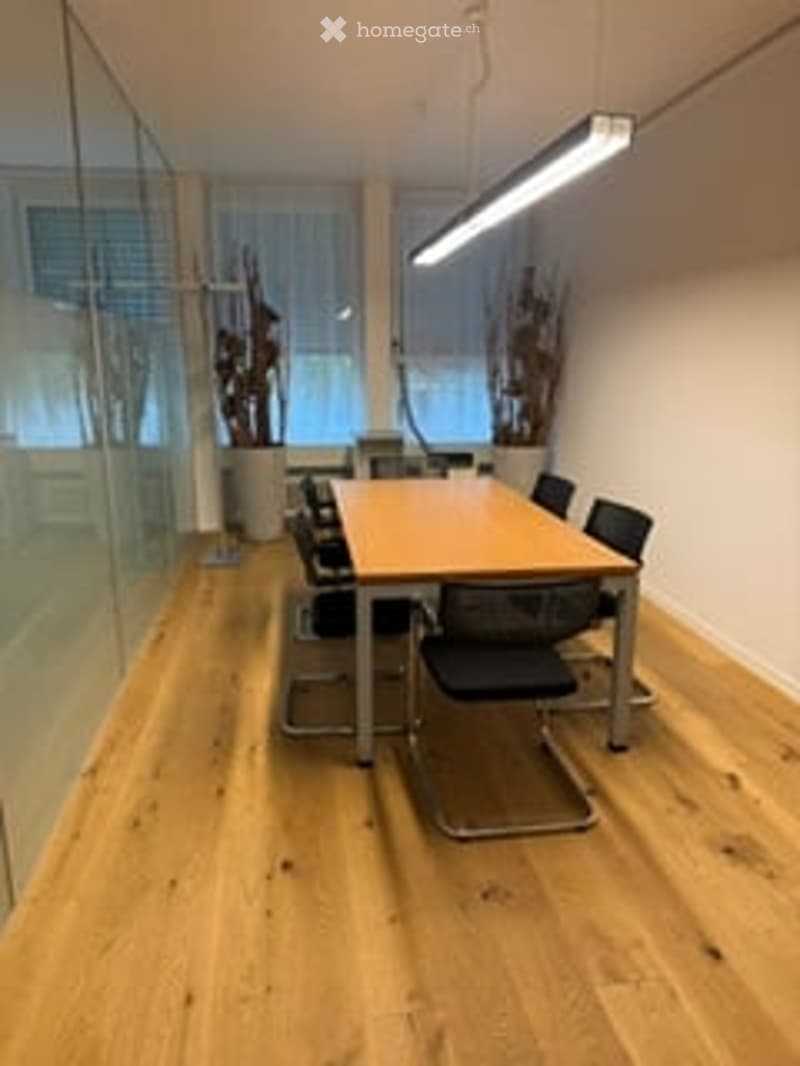 Bright, modern meeting room with a large wooden table and six black chairs, large windows with blinds, and a glass wall.