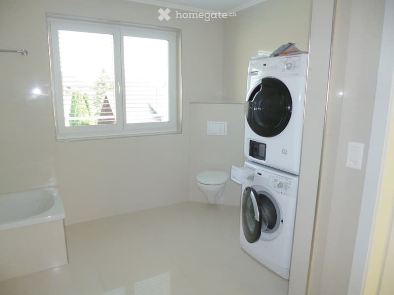 Modern bathroom with a bathtub, toilet, and stacked washing machine and dishwasher unit. The room features light-colored tiles and a large window with blinds.