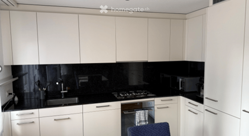 Modern kitchen with white cabinets, black countertops, stainless steel appliances, and a blue chair near the counter.