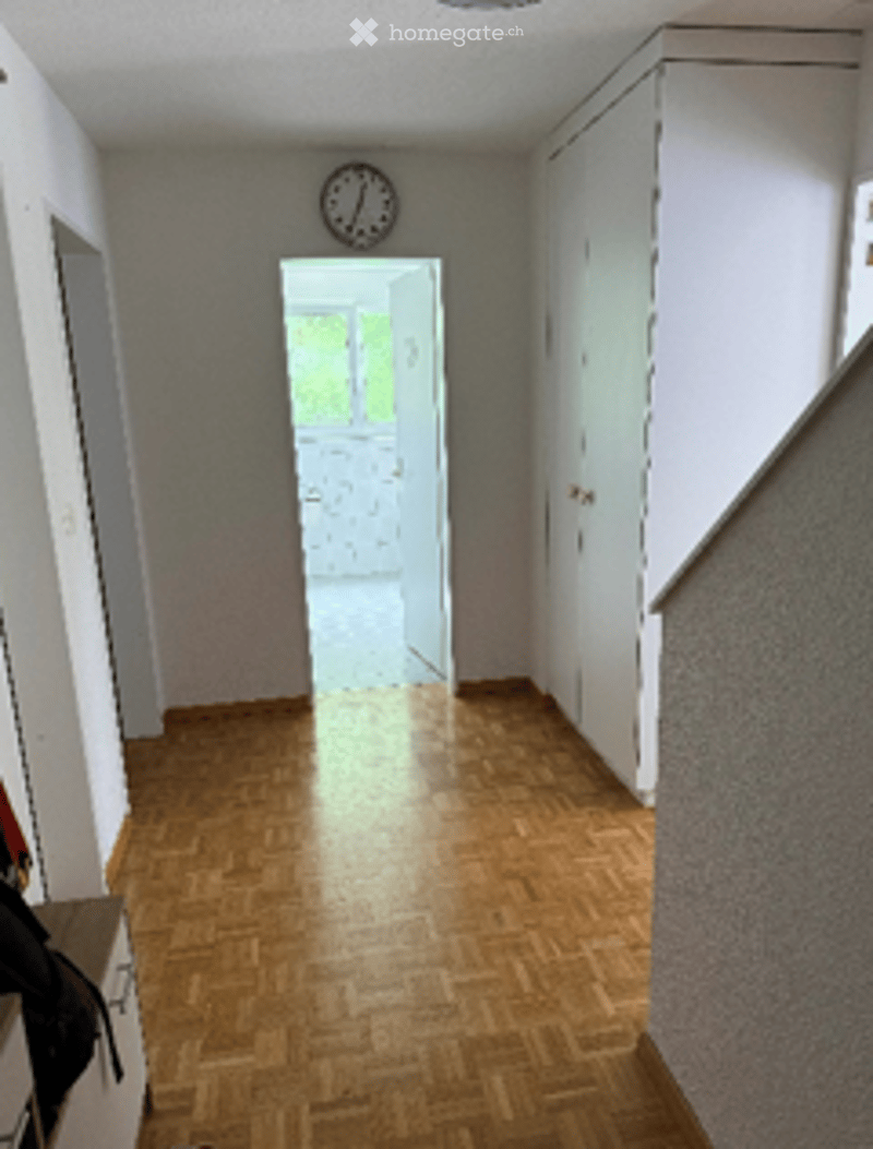 A narrow hallway with wooden flooring, white walls, and a clock on the wall. The hallway leads to an open doorway with natural light coming in.