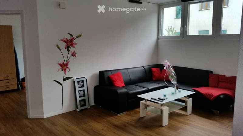 Modern living room with a black leather sofa, red cushions, a white coffee table, a flower vase, and a picture frame on the wall.