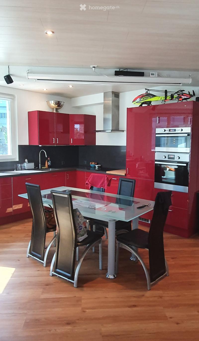 Modern kitchen with red cabinets, stainless steel appliances, a glass dining table with four chairs, and a wooden floor.