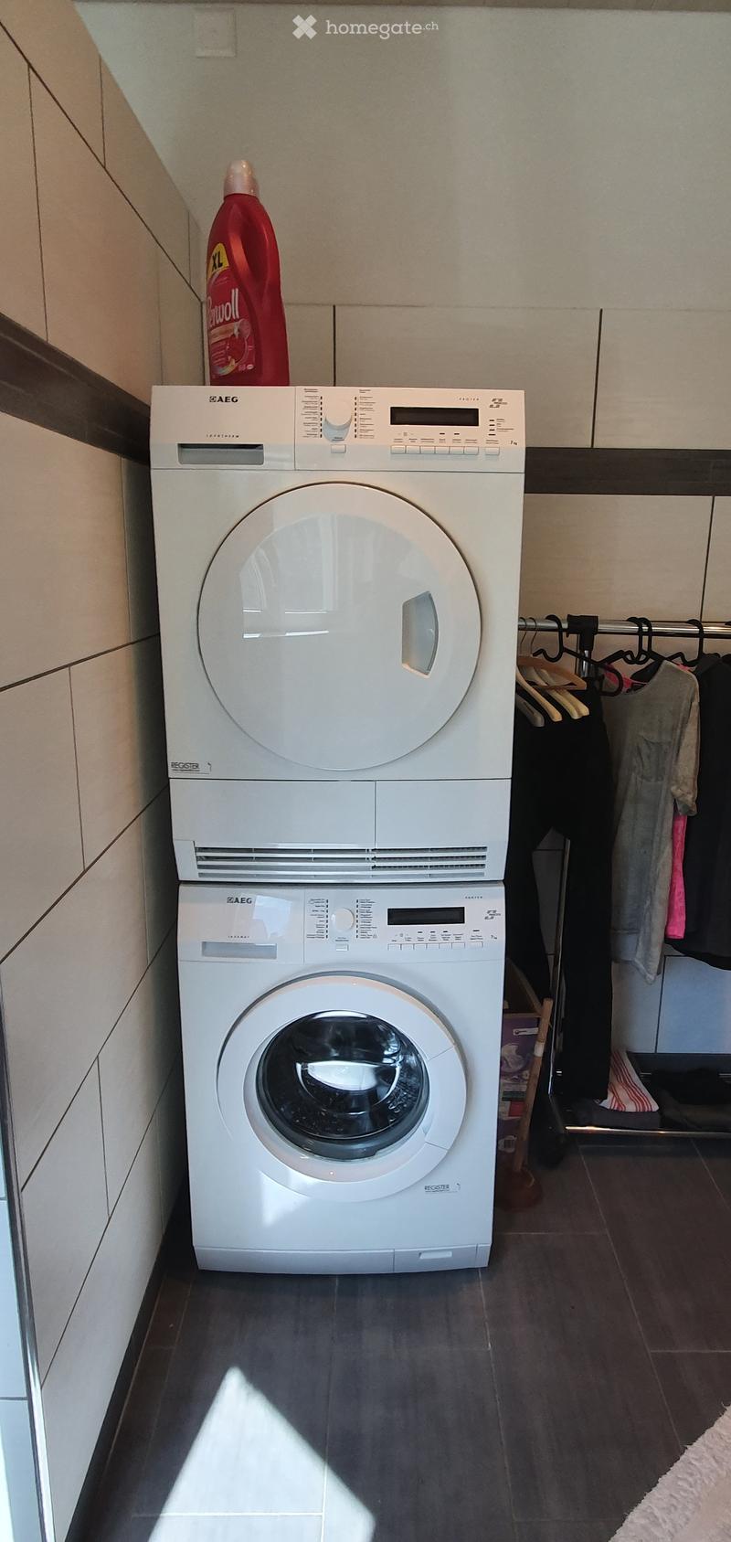 A compact bathroom with a stacked washing machine and dryer unit, a bottle of cleaning solution on top, and a clothes rack with hanging garments.