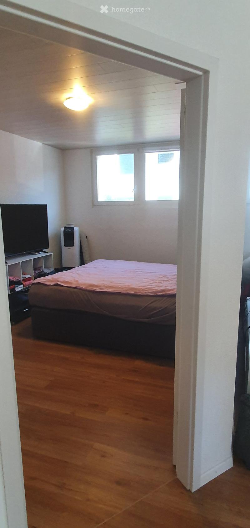 Bedroom with wooden flooring, a bed with a pink blanket, a TV on a white stand, and an air conditioner unit near the window.