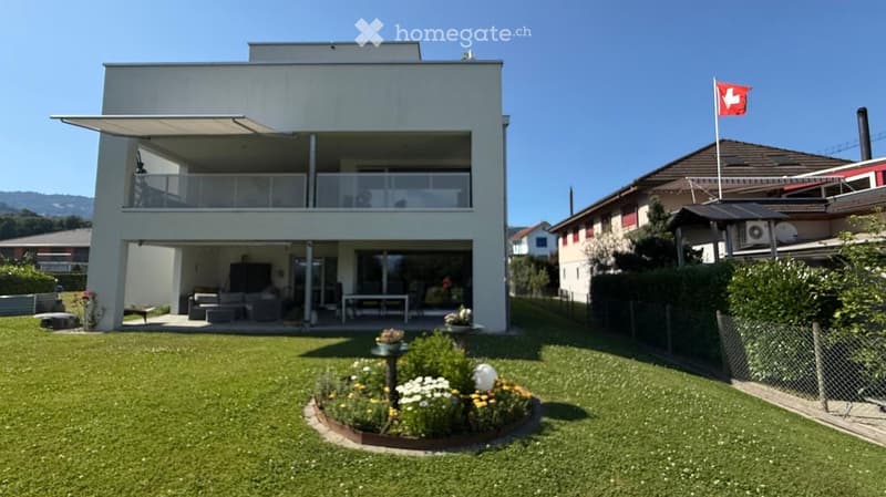 Modern two-story house with a raised ground floor, large windows, and a balcony. The house features a well-maintained lawn, garden, and a Swiss flag on a pole.