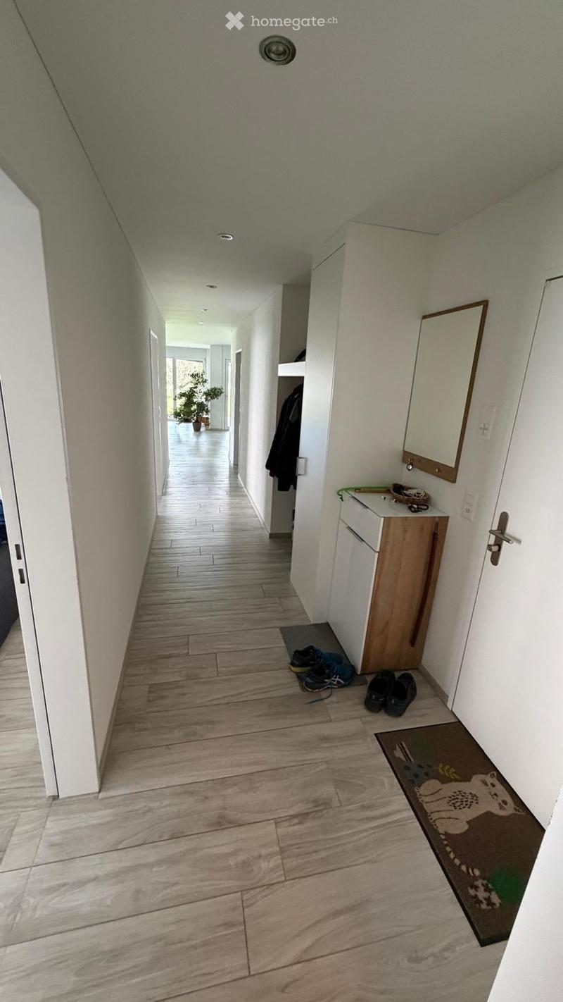A clean, modern hallway with light wooden flooring, white walls, and recessed ceiling lighting. A small wooden cabinet with a mirror and a small rug are visible on the right side, with shoes placed on the floor.