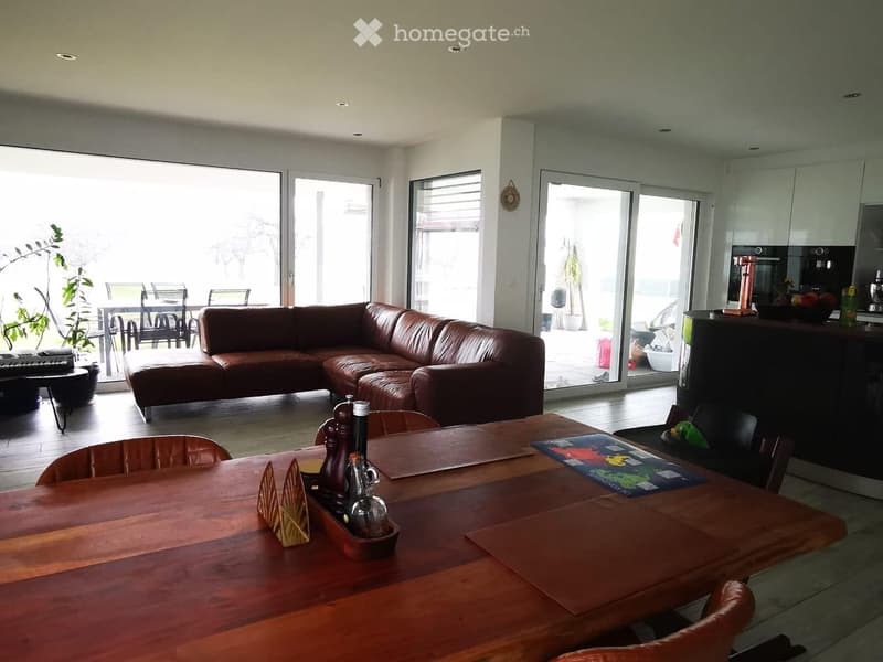 Bright living room with a brown leather sofa, wooden dining table, and kitchen area visible through large glass doors leading to a patio.