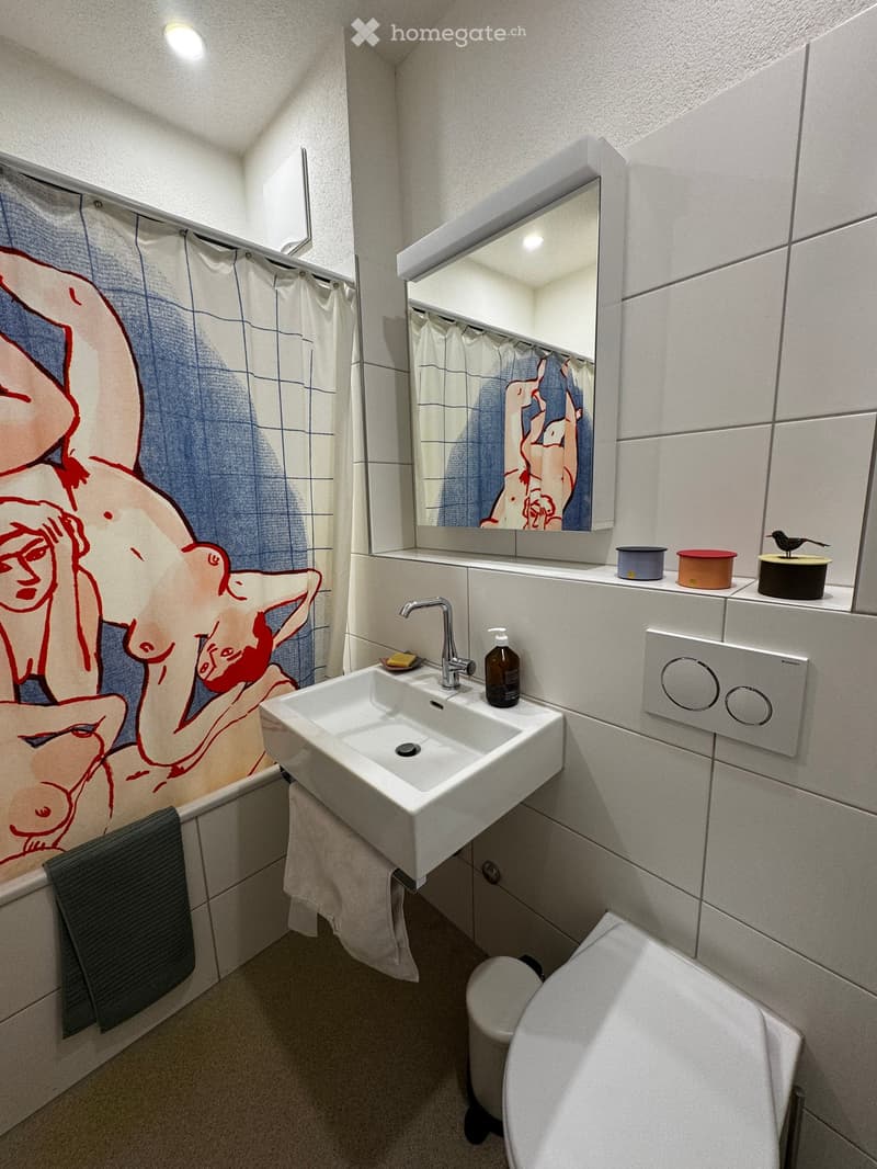 Modern bathroom with white tiles, a sink, mirror, bathtub with a colorful shower curtain, and a small stool. A towel hangs on the side, and decorative items are placed on the counter.