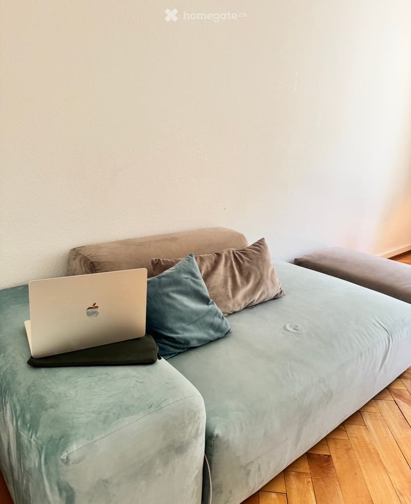 A modern living room with a light green sofa, multiple pillows, and a laptop placed on the sofa.