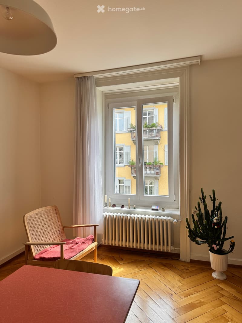 A cozy room with wooden flooring, a wicker chair with a red blanket, a small table, a radiator, and a potted plant near the window.