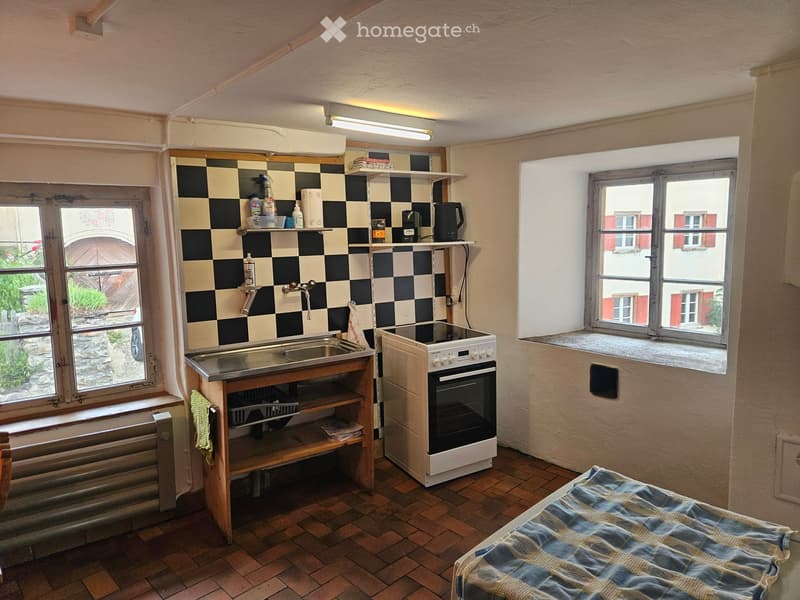 A small kitchen and bedroom combined space with a sink, oven, and a bed. The kitchen area features a checkered backsplash, shelves with cleaning supplies, and a small wooden cabinet. The bedroom area includes a bed with a blue and white blanket.