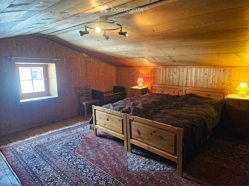Cozy wooden bedroom with slanted ceilings, a single bed with a dark blanket, nightstands with lamps, a wooden chair, and a patterned rug.