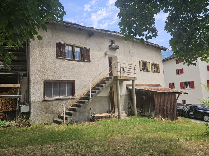 Two-story house with beige exterior, wooden shutters, and a small balcony accessible via exterior stairs. A wooden shed and a stack of firewood are visible to the left, and a car is parked nearby.