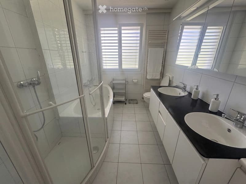 Modern bathroom with a bathtub, double sink vanity, large mirror, and a glass-enclosed shower. The space features tiled walls and floor, a towel rack, and a small cart with toiletries.