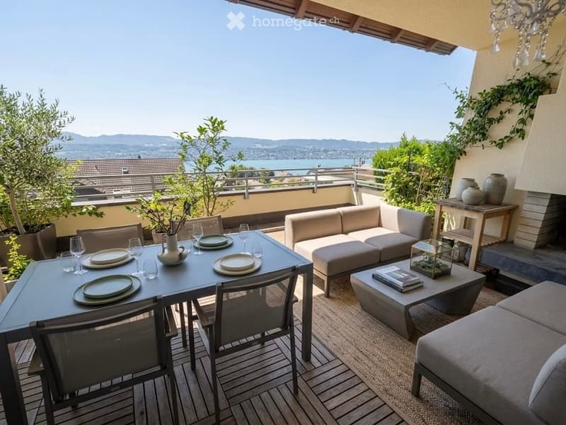Outdoor terrace with a view of a lake and mountains, featuring a dining table with chairs, a sofa, coffee table, and decorative items.