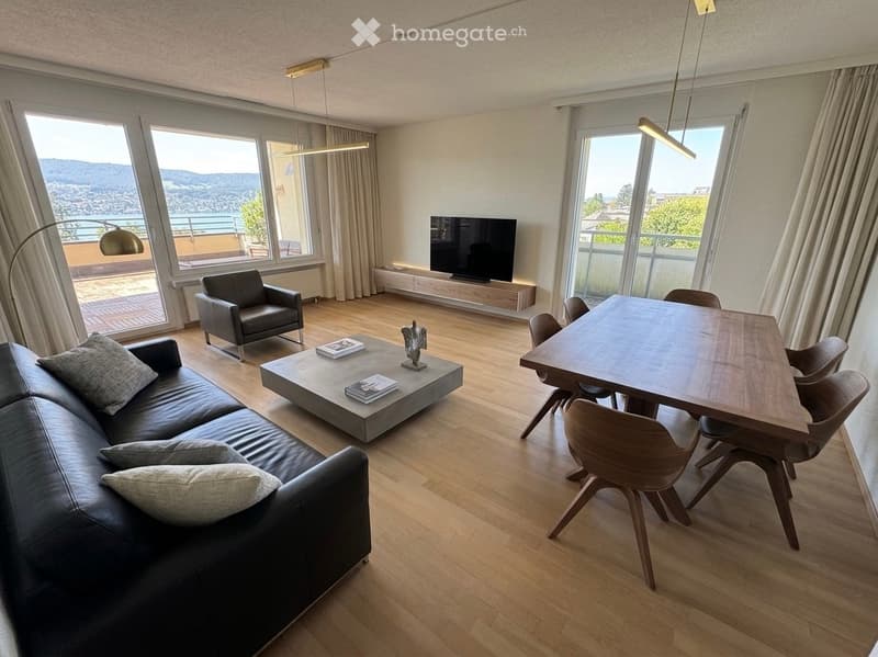 Modern living room with large windows offering a scenic view, featuring a black leather sofa, a wooden coffee table, a dining table with chairs, and a television on a wooden stand.