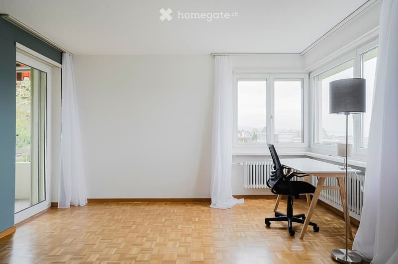 Bright, unfurnished room with wooden flooring, large windows, a desk, a chair, and a floor lamp.