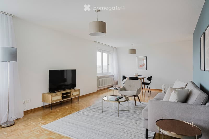 Modern living room with light-colored furniture, a sofa, coffee table, armchair, dining area with chairs and table, television, and floor lamps.