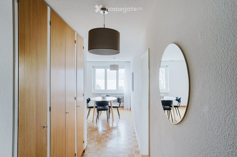 A modern hallway with wooden flooring, white walls, and a large oval mirror on the right side. A round dining table with four chairs is visible in the background, reflected in the mirror.