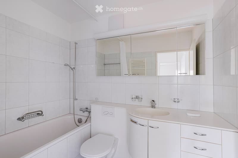 Modern bathroom with white tiles, a bathtub, toilet, sink, mirror, and cabinet.