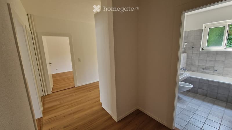 Empty hallway leading to a bathroom with a bathtub, toilet, and tiled walls. The hallway has wooden flooring and white walls.