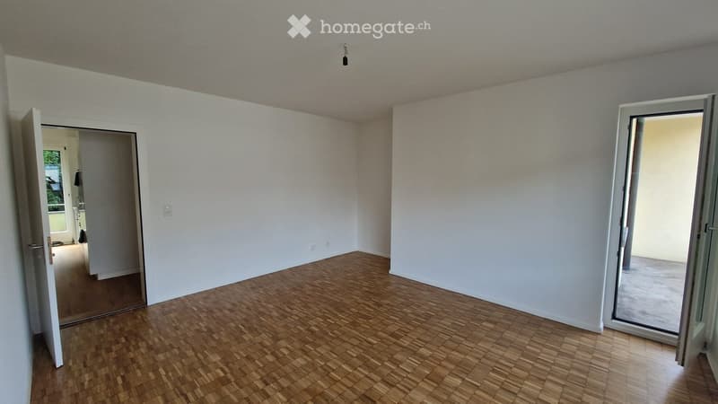 Empty room with wooden flooring, white walls, and large sliding glass door leading outside. An open doorway on the left reveals another room.