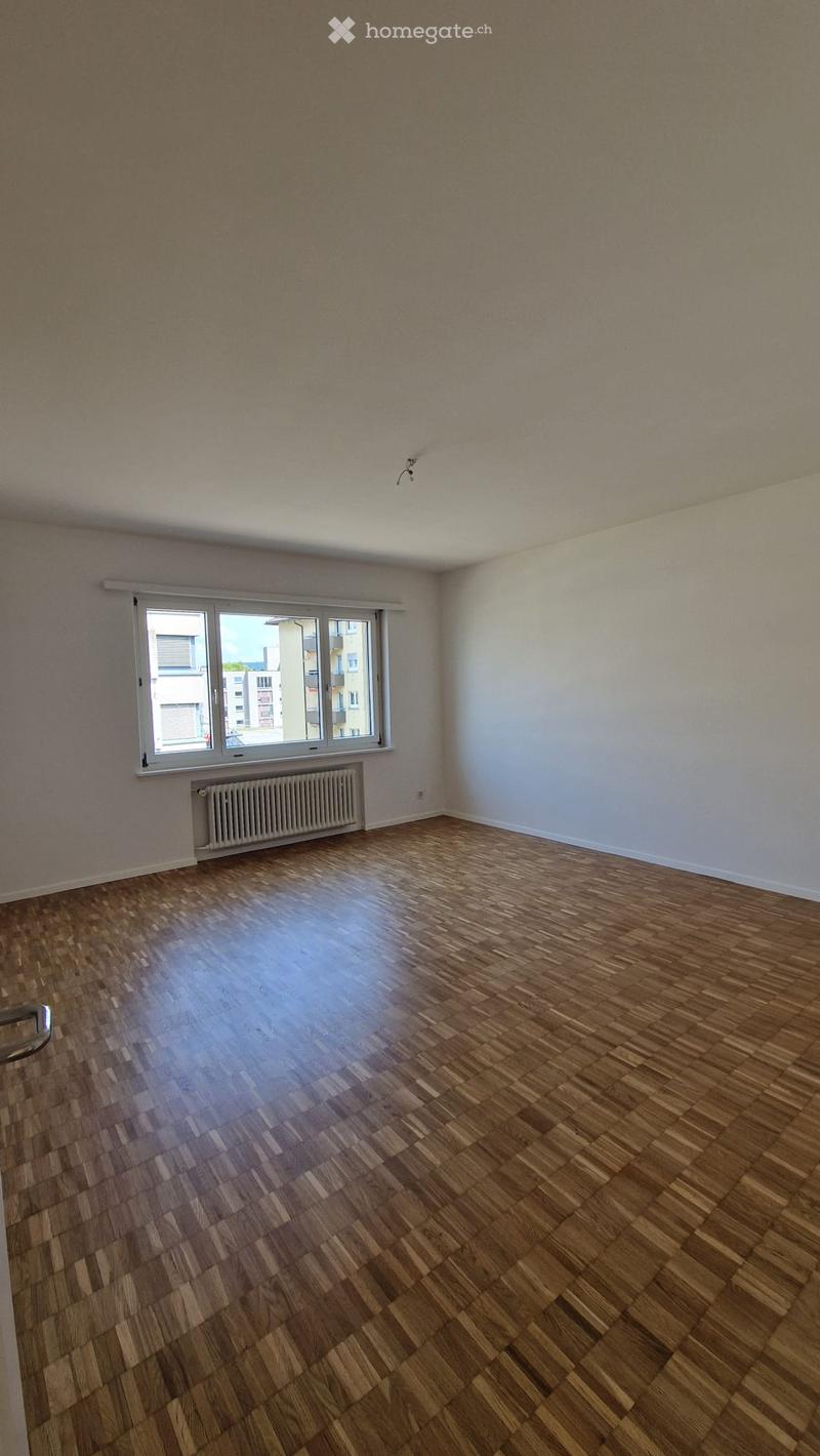 Empty room with wooden flooring, white walls, a large window with white frame, and a radiator below the window.