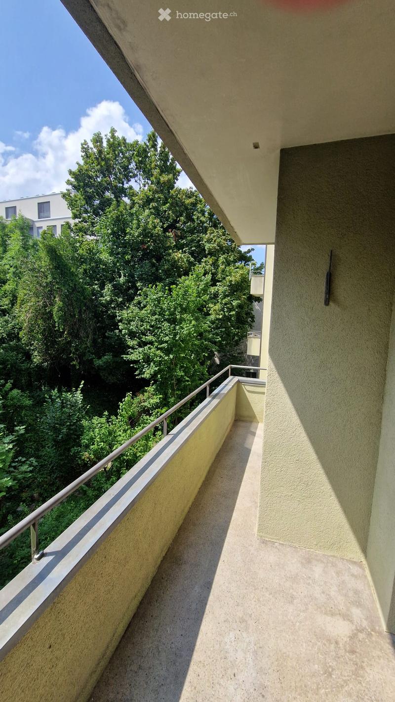 Balcony with metal railing, overlooking a green area with trees and a building in the background.