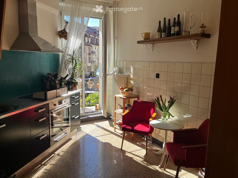 A small, well-lit kitchen with modern appliances, a red chair, a small table, and shelves with bottles and a vase.