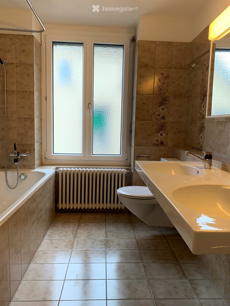 Modern bathroom with beige tiles, a bathtub, a toilet, a double sink, and a radiator. The room features large windows with frosted glass and a shower area with a glass door.