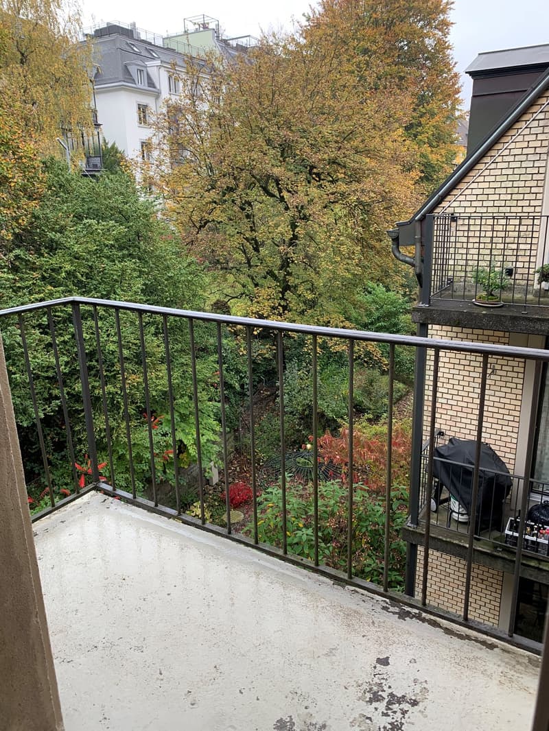 Balcony with metal railing, overlooking a lush green garden area with trees and shrubs. The balcony floor is concrete, and there is a small covered area with a grill and some storage items on the right side.
