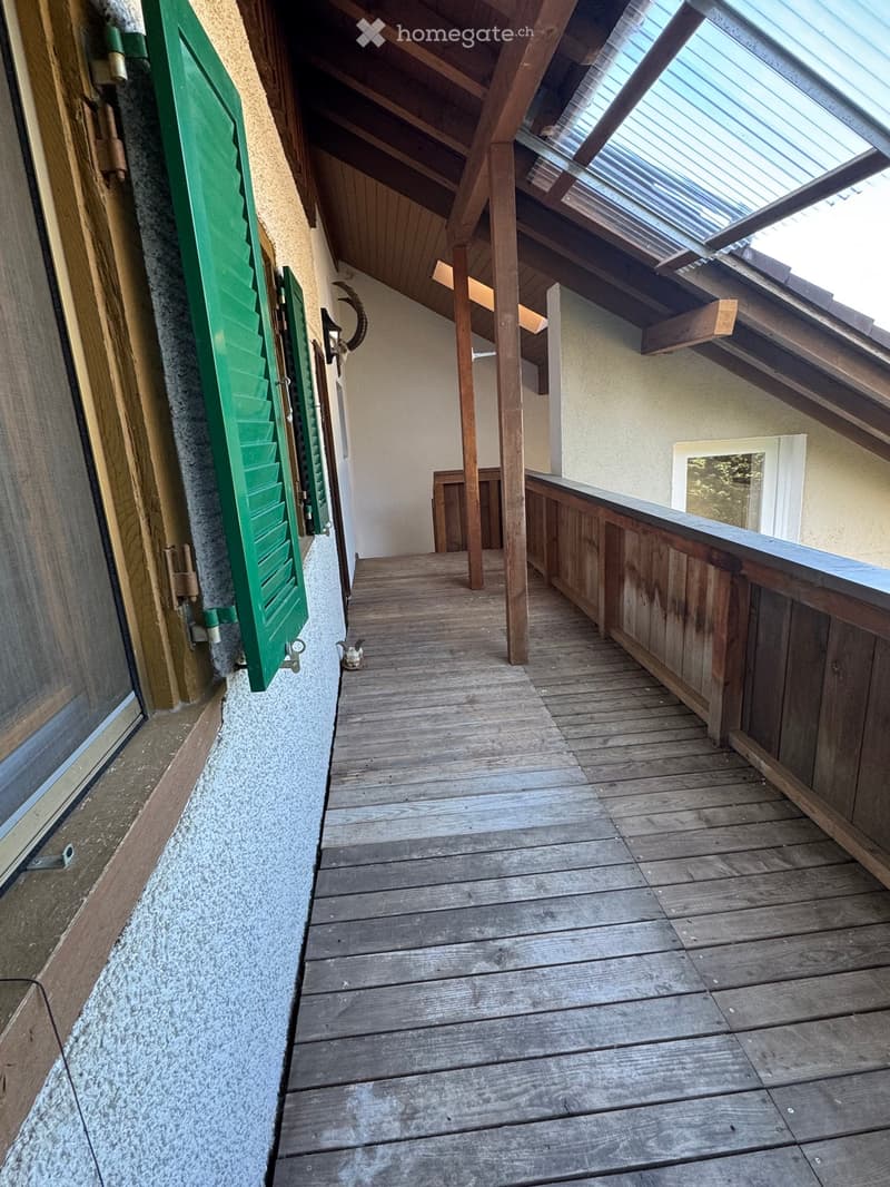 Wooden balcony with slanted wooden ceiling, green shutters, and a small window at the end.