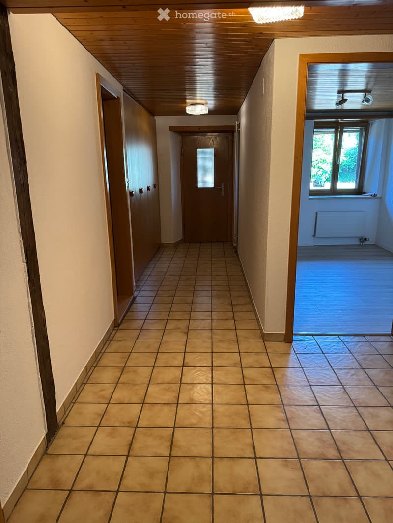 Empty hallway with tiled floor, wooden ceiling, and several doors leading to other rooms. Natural light from a window on the right side.