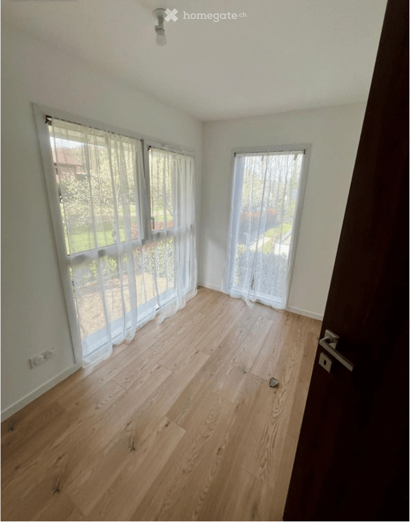 Empty room with light wooden flooring, white walls, large windows with sheer curtains, and a door visible on the right side.