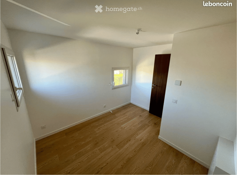 Empty room with wooden flooring, white walls, and two windows, one of which is partially open. A closed wooden door is visible on the right side.