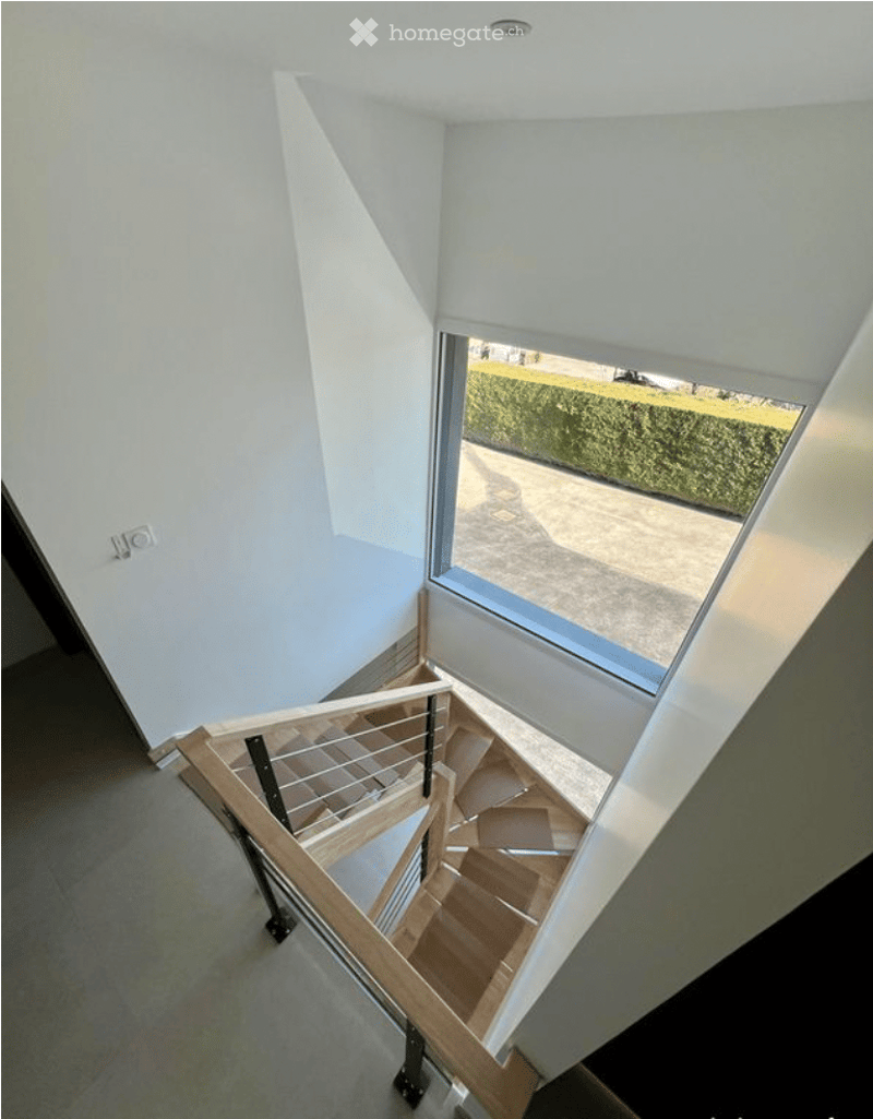 Modern staircase with wooden steps and metal railing, leading to an upper level. The walls are painted white, and there is a large window at the top providing natural light and a view of the garden outside.