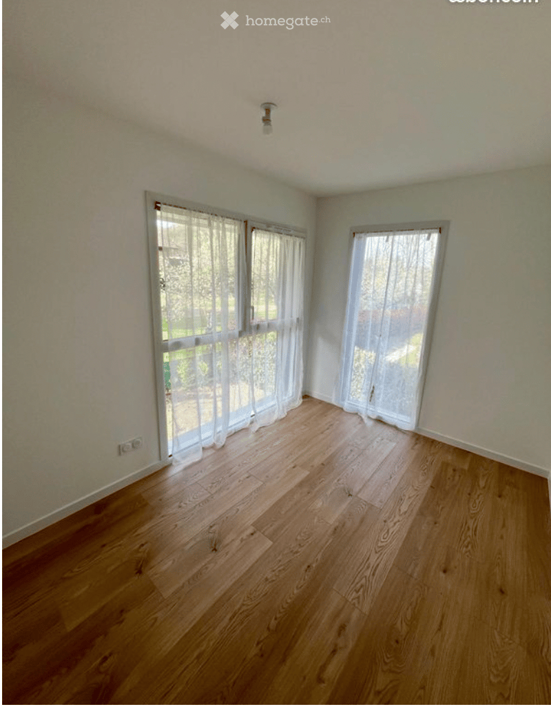 Empty room with wooden flooring, white walls, and large windows with sheer curtains. No furniture or equipment present.