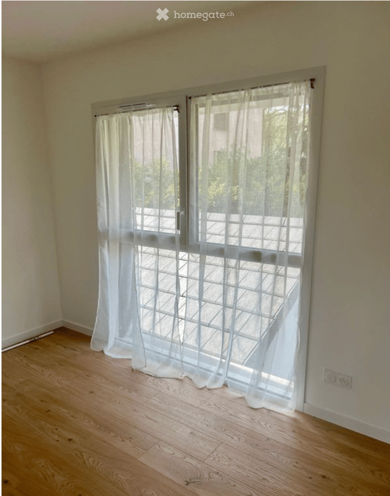 Empty room with wooden flooring, large window with white curtains, and a view of a balcony area.