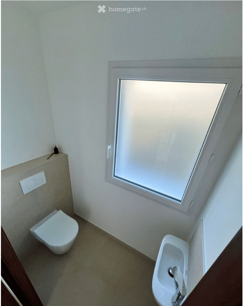 Compact bathroom with a toilet, bidet, and a small window for natural light. The walls are painted white, and the floor is tiled.