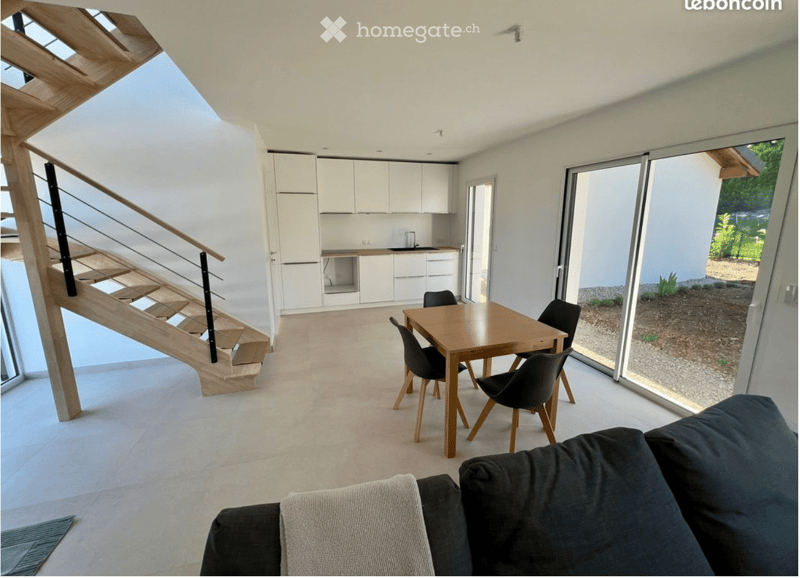 Open-plan living and dining area with a modern sofa, wooden dining table, chairs, and white kitchen cabinets.