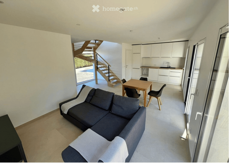 Open-plan living and dining area with a dark blue sectional sofa, a wooden dining table with chairs, and a modern white kitchen area with cabinets and a sink.