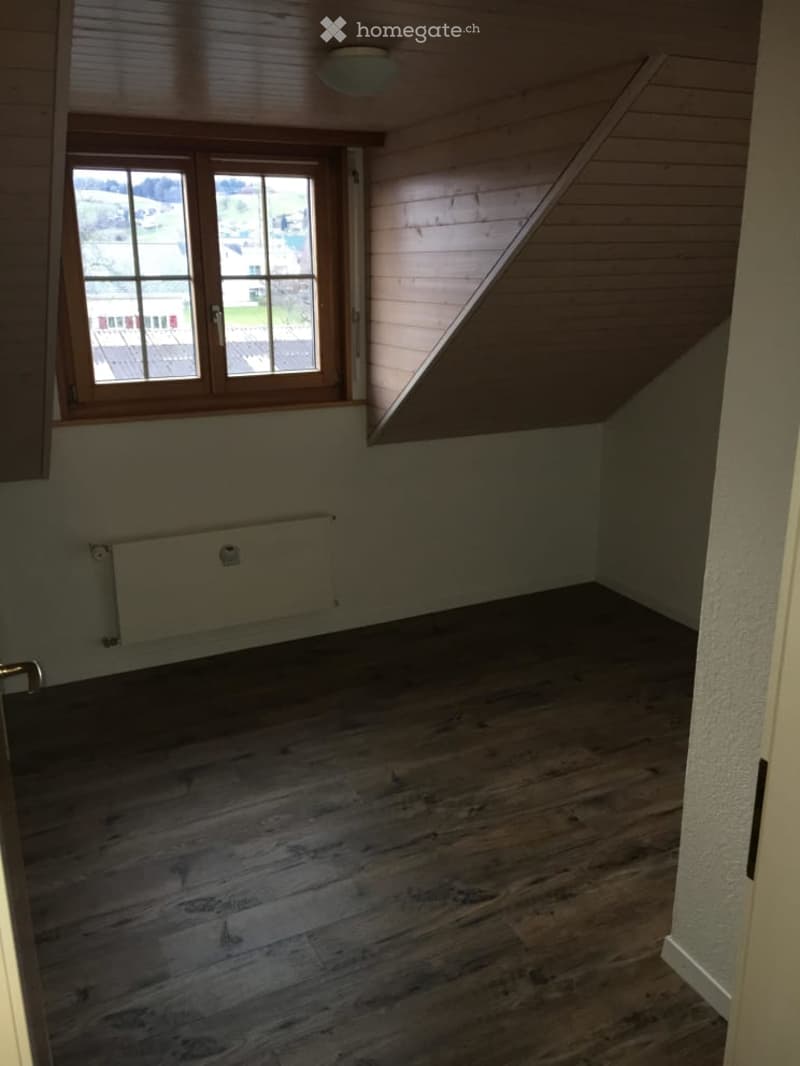 Empty attic room with wooden slanted ceiling, wooden floor, large window, radiator, and visible light fixture.