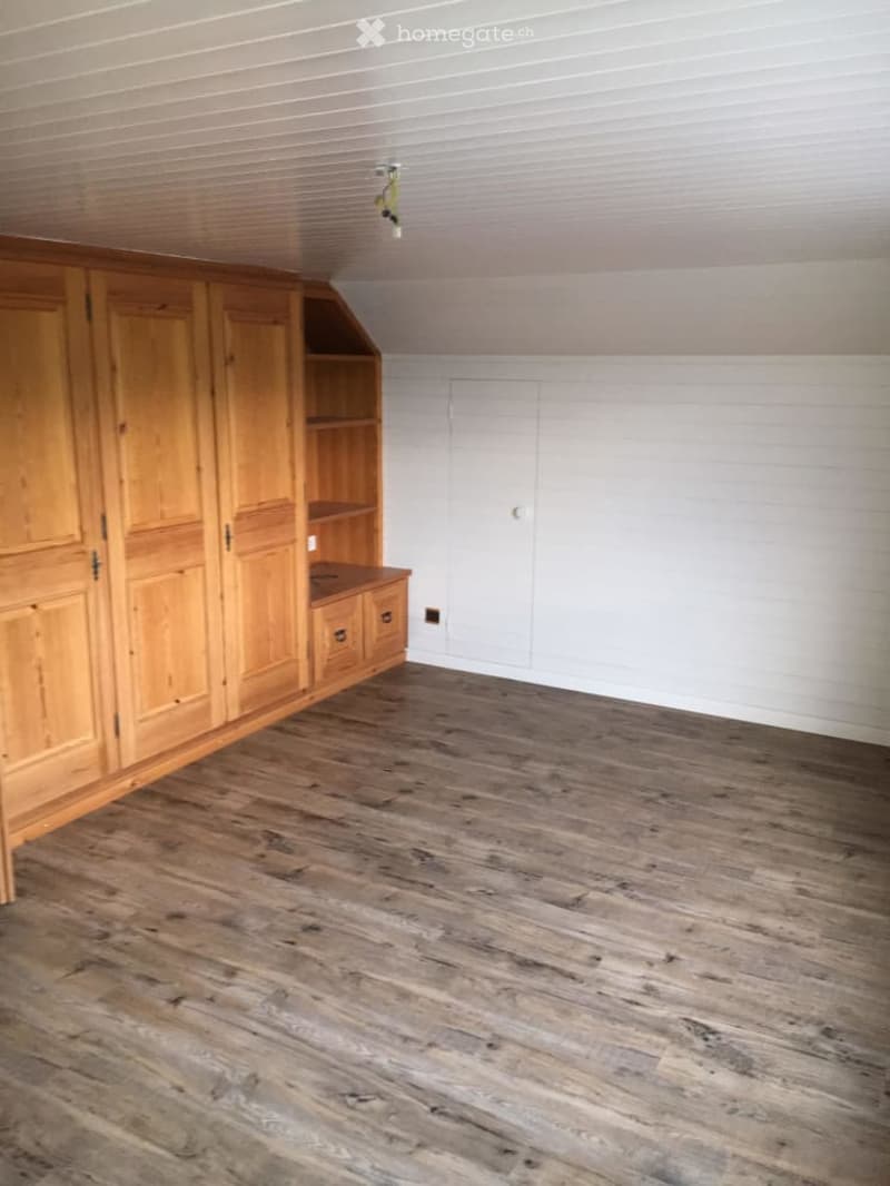Empty room with wooden flooring and white walls, featuring a large wooden wardrobe with shelves and drawers.
