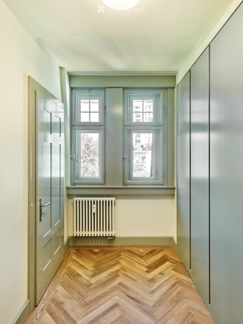 A narrow hallway with herringbone-patterned wooden flooring, light-colored walls, and a radiator below a large window. The hallway features a closed door on the left and a window with multiple panes on the right.
