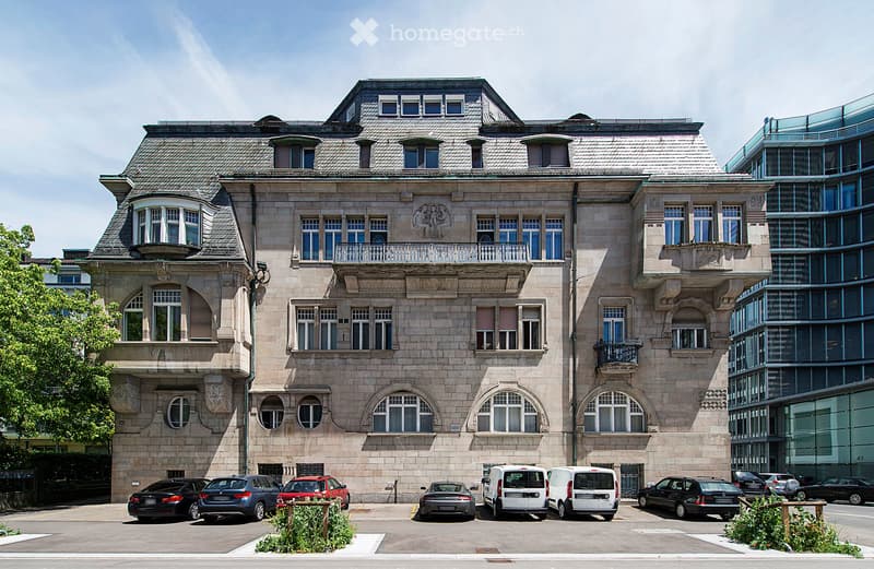 A large, multi-story building with a classic architectural style, featuring arched windows, decorative stonework, and a prominent roofline. The building has several balconies and is surrounded by parked cars and greenery.