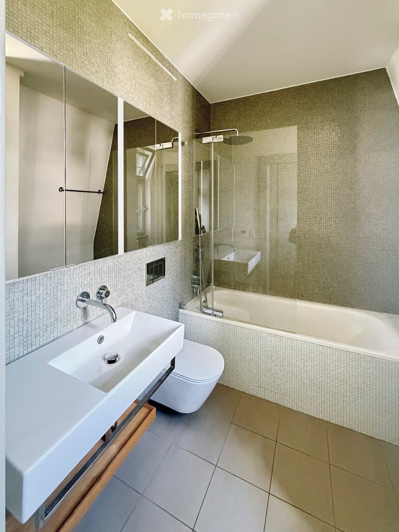 Modern bathroom with a white sink, toilet, bathtub, and large mirror. Features tiled walls and a glass-enclosed shower area.