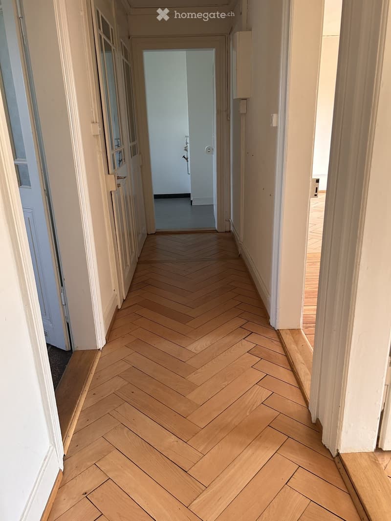 Hallway with herringbone-patterned wooden flooring, white walls, and doors on both sides.