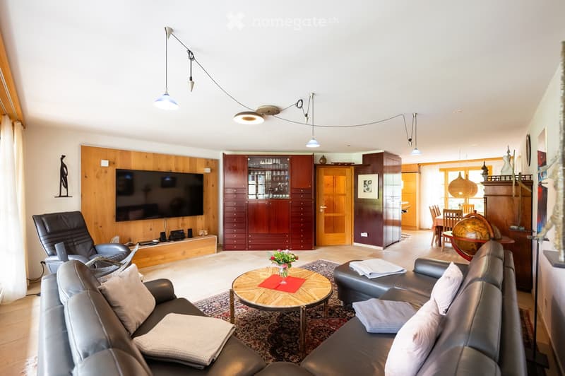 Modern living room with a large sectional sofa, coffee table, armchair, TV screen, and a dining area visible in the background.