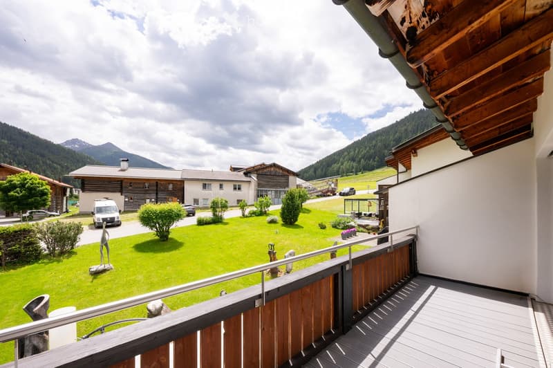 Balcony overlooking a green lawn with trees, sculptures, and a view of a village with houses and mountains in the background.