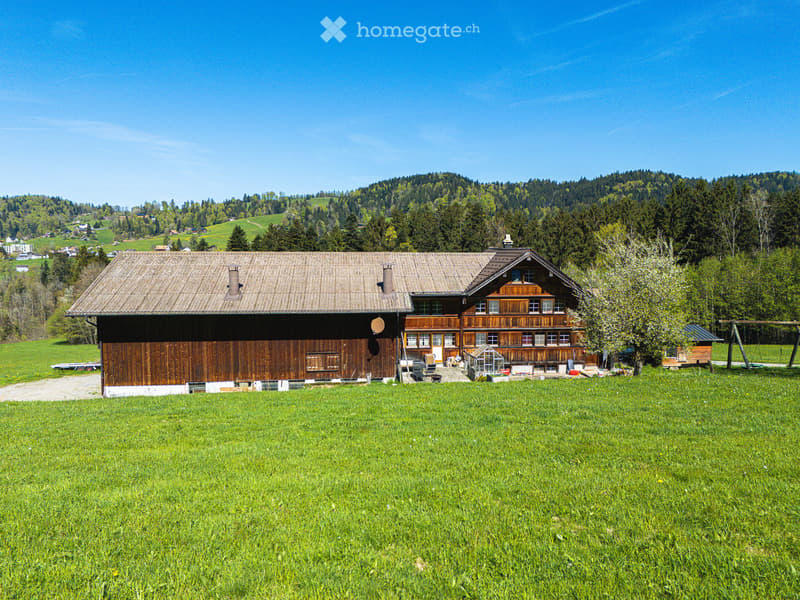 Traditional wooden house with a sloped roof, surrounded by green fields and forested hills under a clear blue sky.